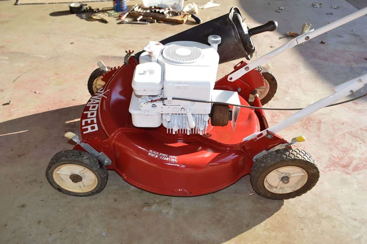 An old red walk-behind petrol mower with a white engine standing on a concrete driveway