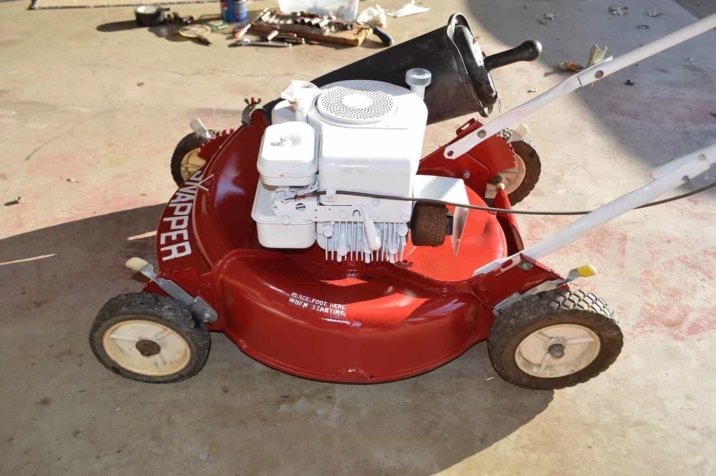 An old red walk-behind petrol mower with a white engine standing on a concrete driveway