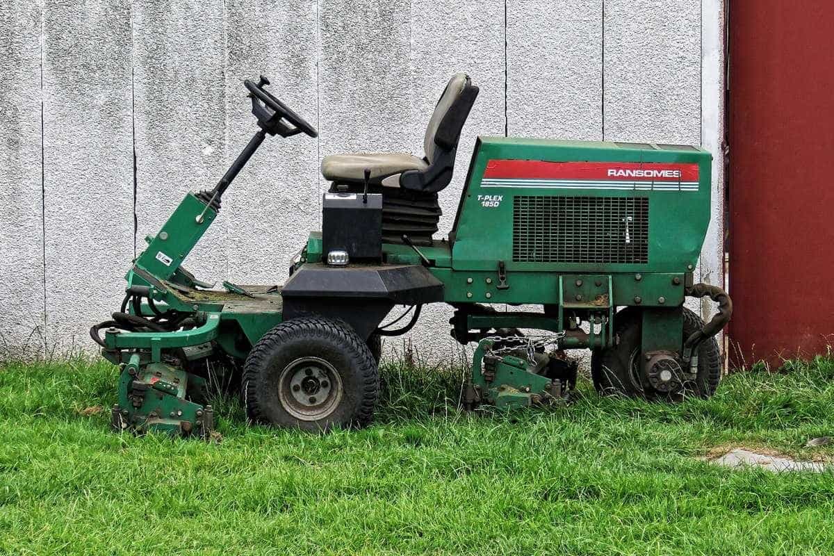 A green ride-on reel mower parked on grass beside a white weatherboard building